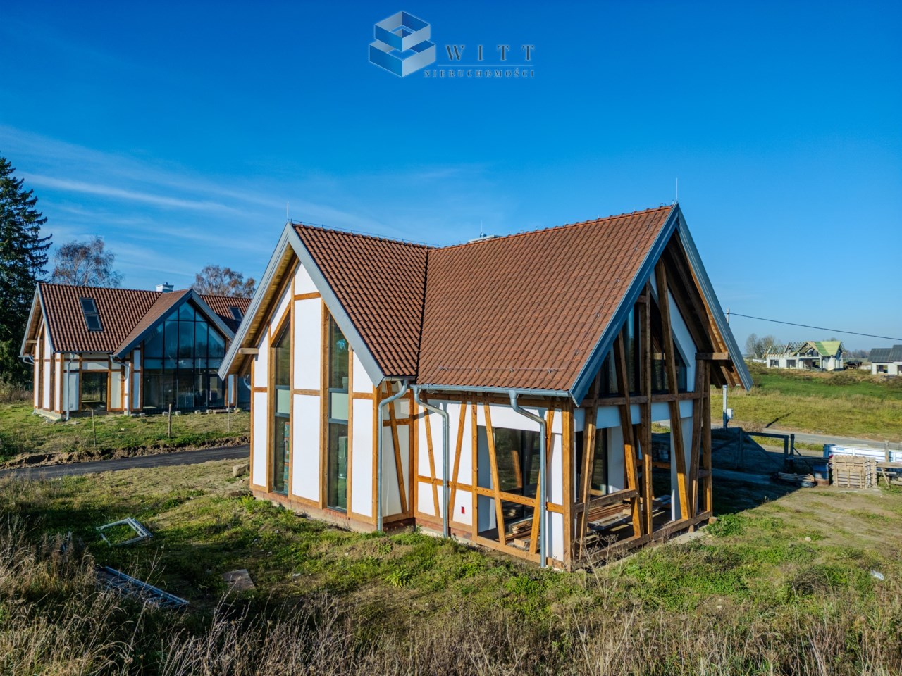 Nowoczesny dom w stylu ekologicznym z drewnianymi elementami, otoczony zielenią, na tle błękitnego nieba. Widać sąsiednie budynki oraz przestrzeń do zagospodarowania.