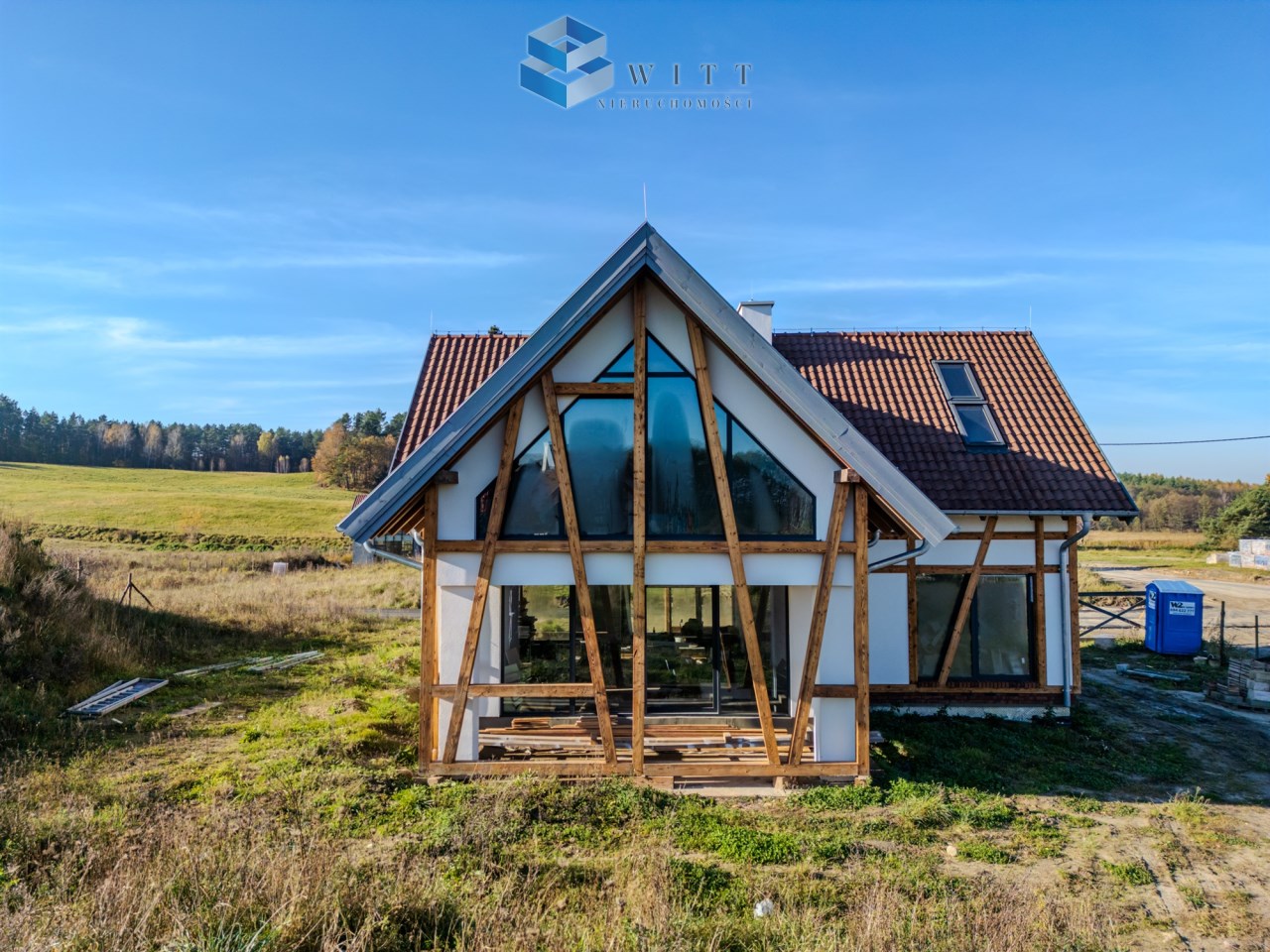 Nowoczesny dom jednorodzinny z dużymi oknami i drewnianymi elementami konstrukcyjnymi, otoczony zielonym terenem.