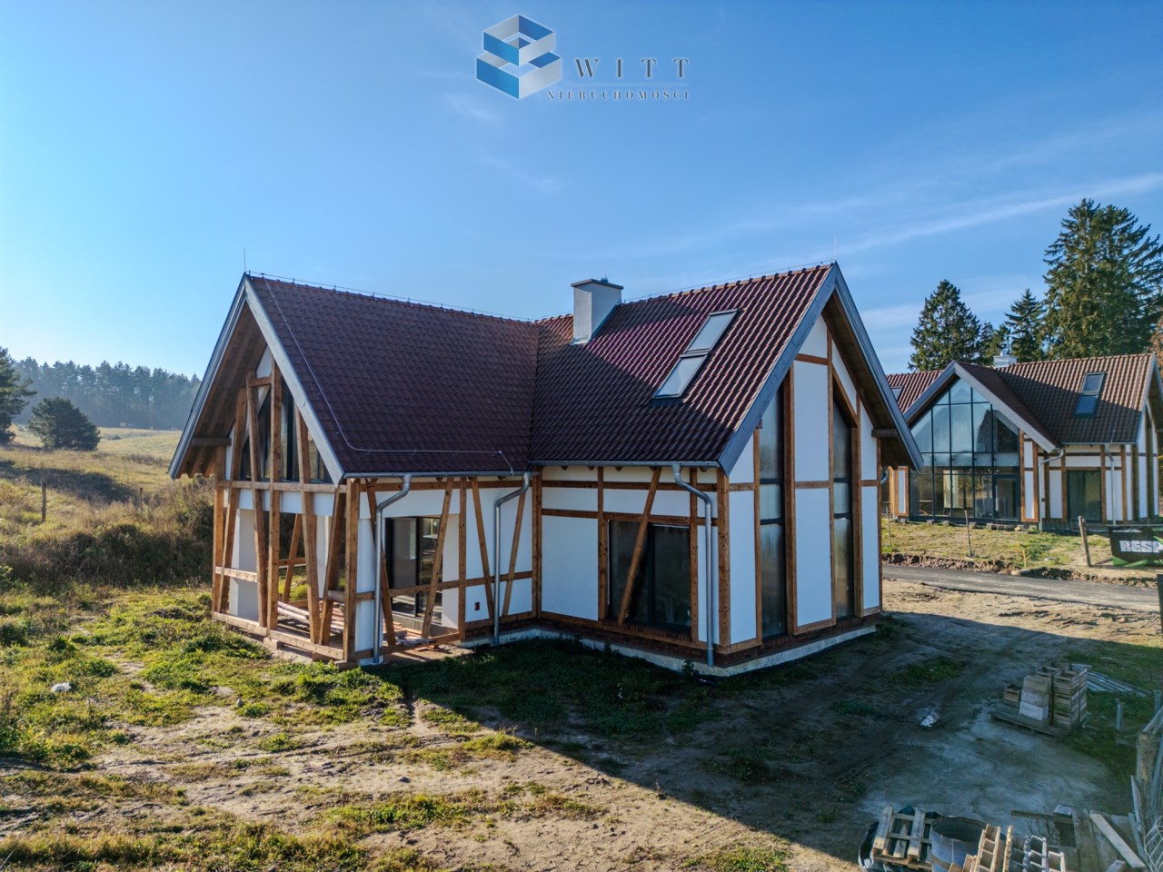 Nowoczesny dom w stylu szkieletowym z czerwonym dachem, otoczony zielenią i drzewami. Widok z boku na budynek z dużymi oknami i drewnianymi elementami konstrukcyjnymi.