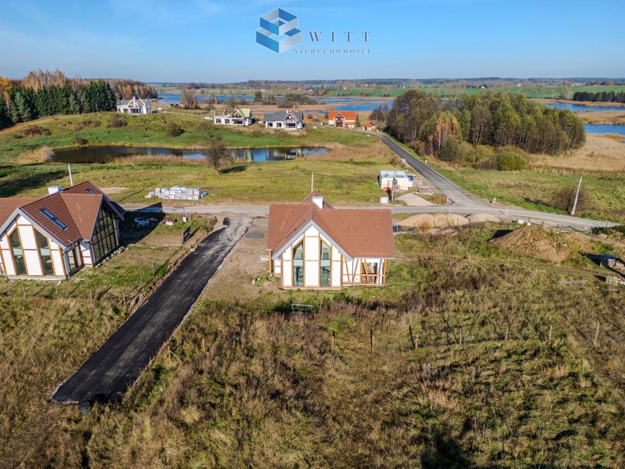 Widok z lotu ptaka na nowoczesne domy w malowniczej okolicy, z drogą, stawem i terenami zielonymi.
