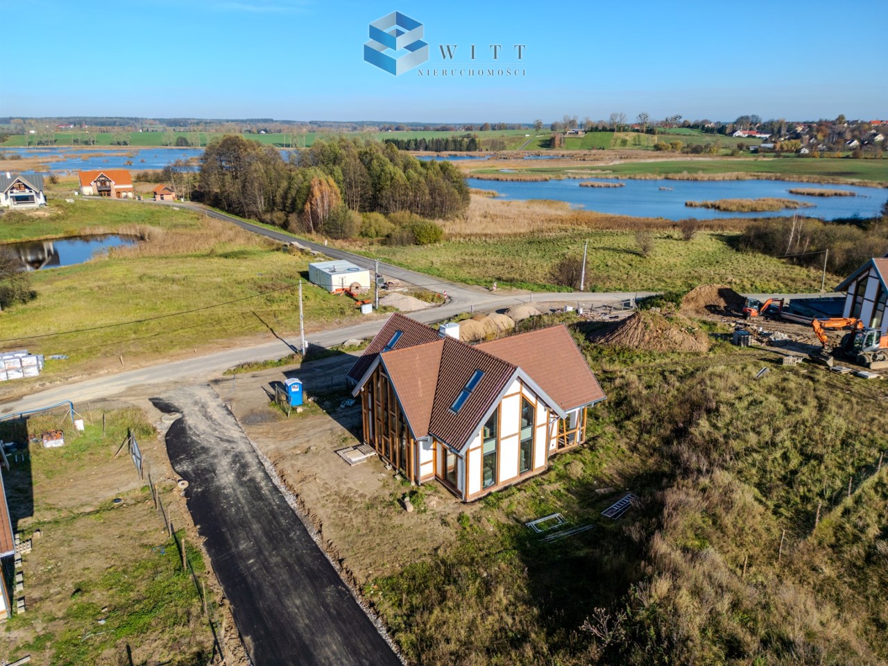 Widok z lotu ptaka na nowoczesny dom z poddaszem, otoczony zielenią, w pobliżu jeziora i drogi. W tle widać inne budynki oraz tereny budowlane.