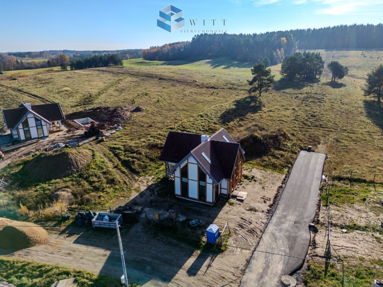 Widok na nowoczesny dom w trakcie budowy w otoczeniu zieleni, z drogą dojazdową i sprzętem budowlanym w tle.