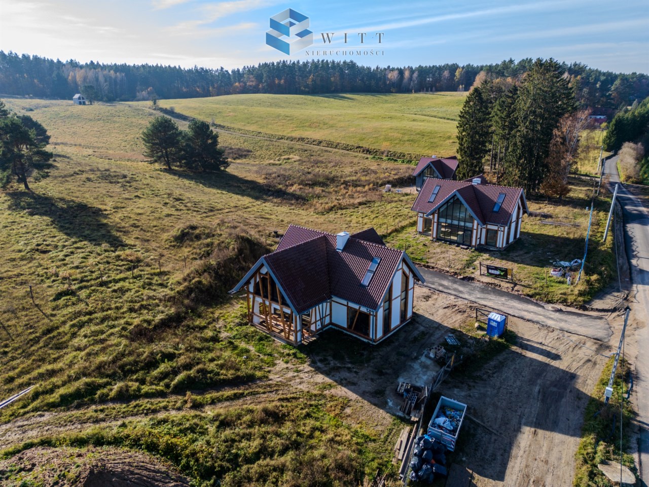 Widok na nowoczesne domy w stylu wiejskim w otoczeniu zielonych pól i lasów, z drogi prowadzącej na obrzeżach miejscowości.