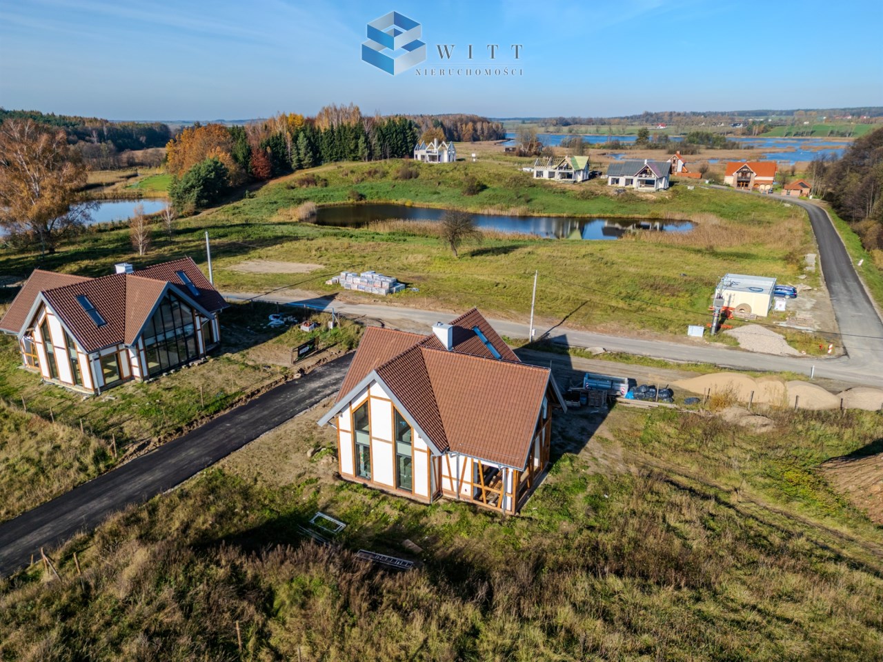 Widok z lotu ptaka na nowe domy w malowniczej okolicy, z zielonymi terenami i stawem w tle. Widać także drogę oraz budowę. Idealne miejsce dla osób szukających spokojnego życia na wsi.