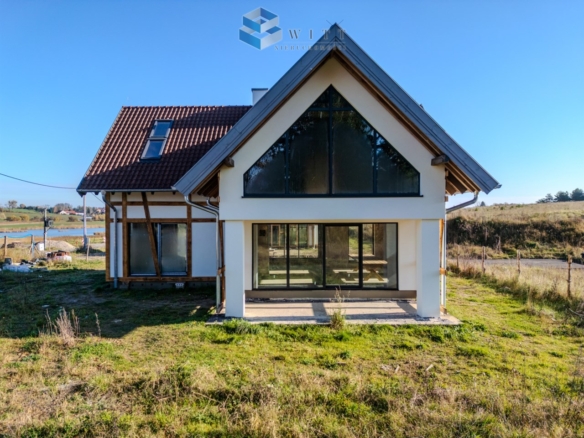 Nowoczesny dom jednorodzinny z dużymi oknami, położony w malowniczej okolicy, z widokiem na tereny zielone i wodę.