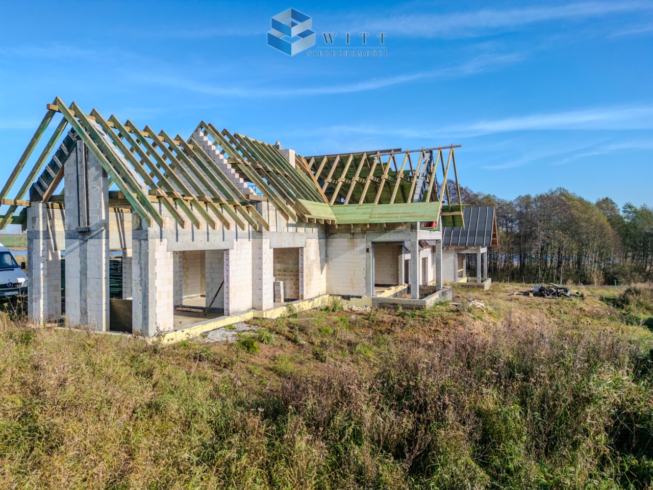 Budowa domu jednorodzinnego w stanie surowym, z widocznymi konstrukcją dachu i ścianami z bloczków. Teren otoczony zielenią, w tle drzewa i błękitne niebo.