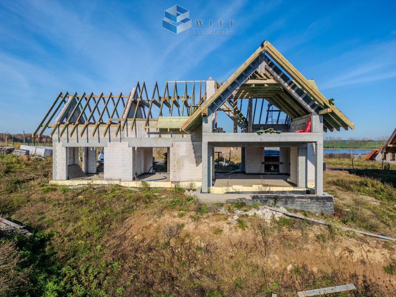 Budowa nowoczesnego domu jednorodzinnego w stanie surowym, z widoczną konstrukcją dachu i ścianami z bloczków betonowych, otoczona naturalnym terenem.