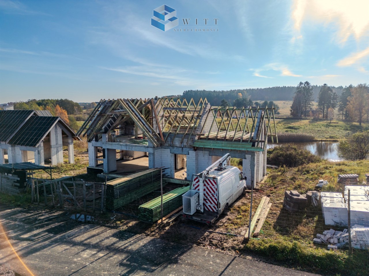 Budowa domu z widokiem na teren zielony, z widoczną konstrukcją dachu oraz materiałami budowlanymi w otoczeniu. Na pierwszym planie znajduje się furgonetka budowlana.