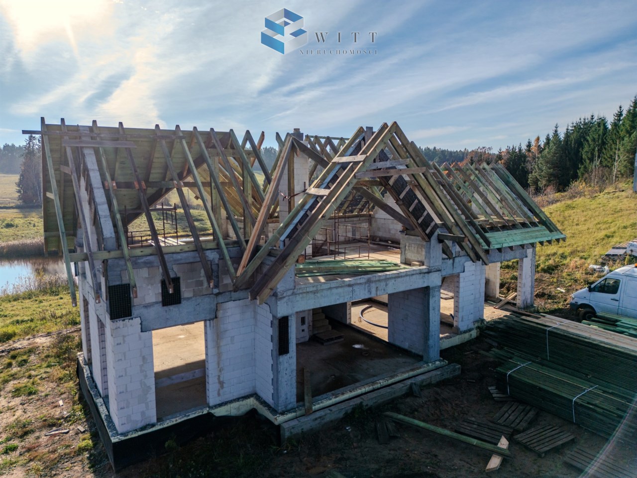 Rozpoczęta budowa domu jednorodzinnego z drewnianym dachem w trakcie konstrukcji, otoczenie zieleni i naturalnego krajobrazu.