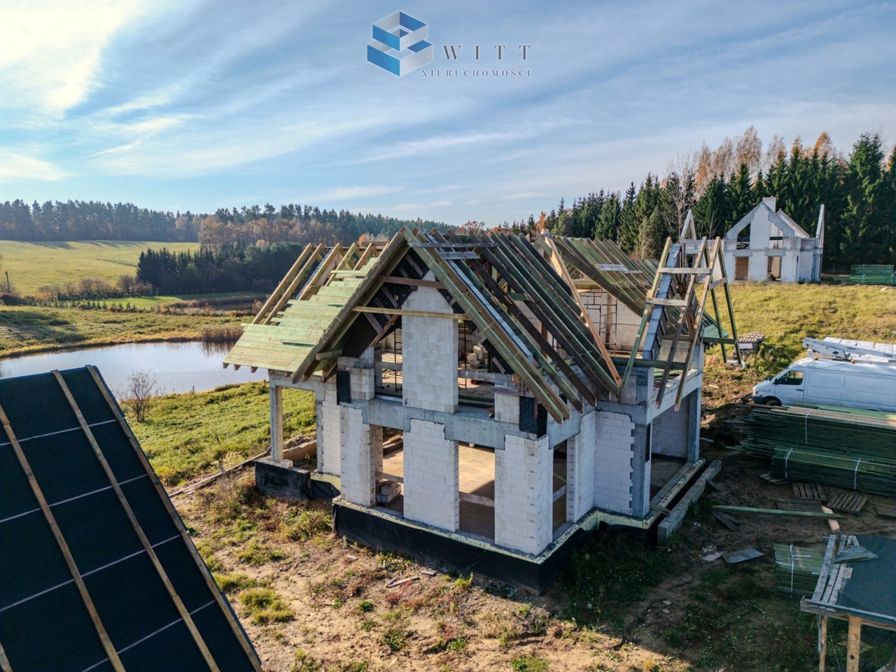 Budowa domu jednorodzinnego w otoczeniu natury, z widokiem na staw i las. Widać konstrukcję dachu oraz materiały budowlane na placu.