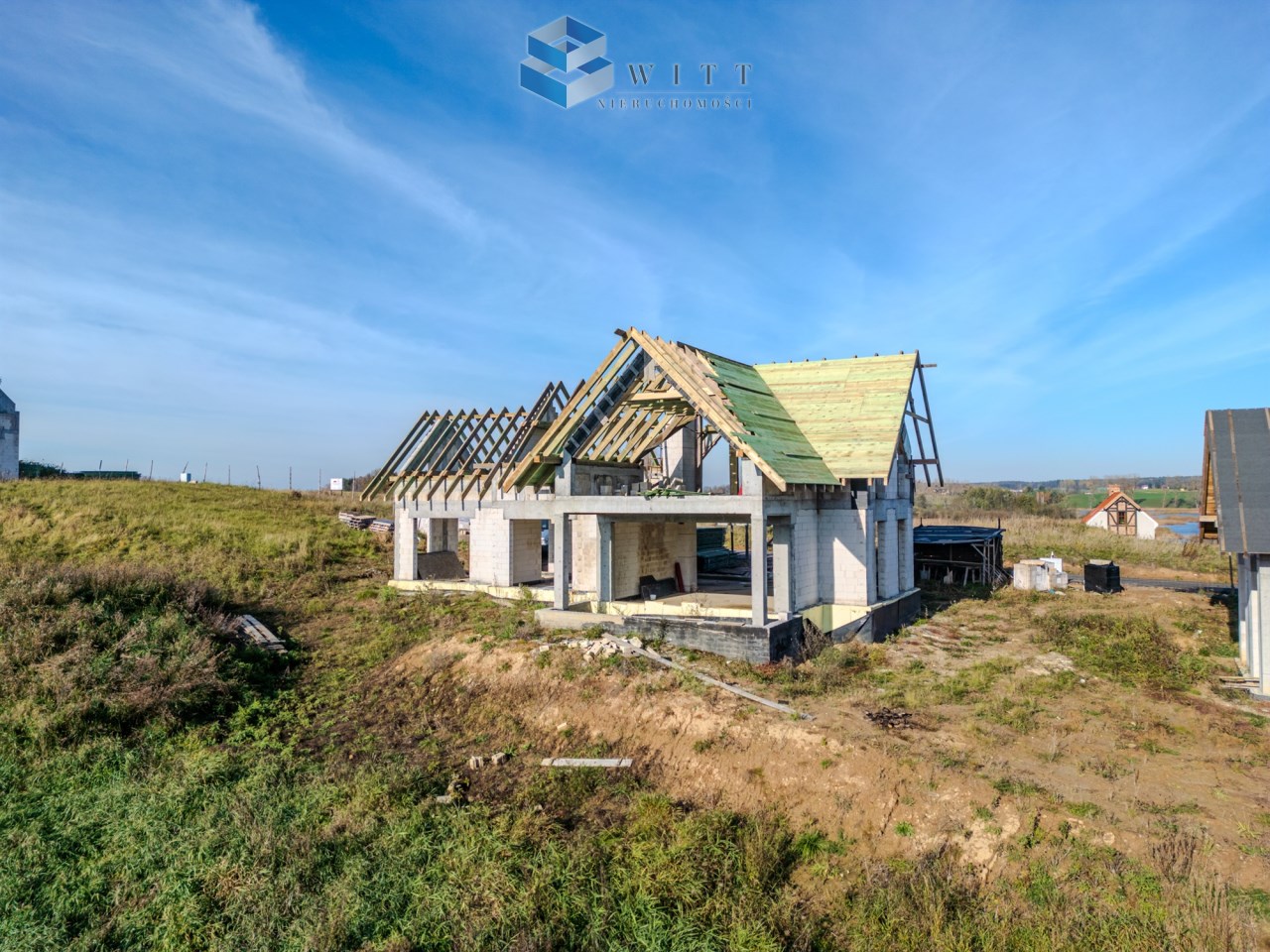 Budowa domu jednorodzinnego w trakcie realizacji, widok na nieukończoną konstrukcję z drewnianym dachem, otoczenie zielonych łąk i innych budynków mieszkalnych.