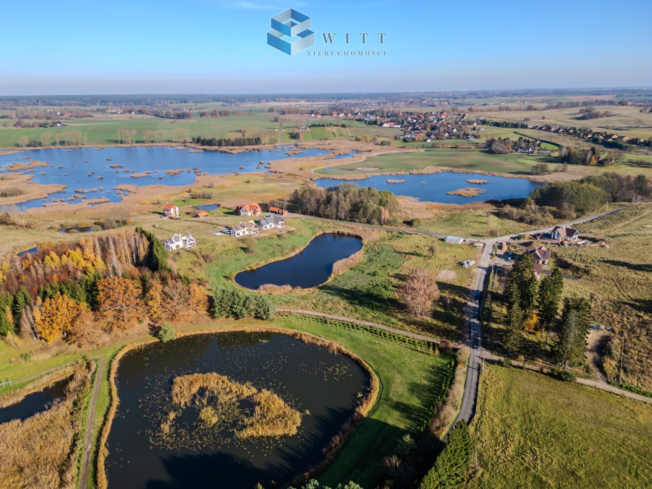 Widok z lotu ptaka na malowniczy krajobraz z jeziorami, stawami i terenami zielonymi, z zabudowaniami mieszkalnymi w tle. Idealne miejsce do wypoczynku i aktywności na świeżym powietrzu.