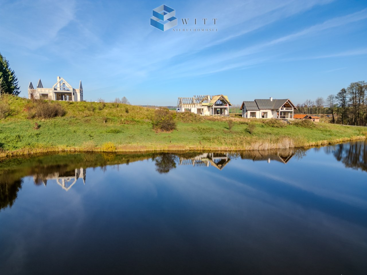 Widok na nowoczesne domy nad spokojnym stawem, otoczone zielonymi wzgórzami i drzewami, w słoneczny dzień.