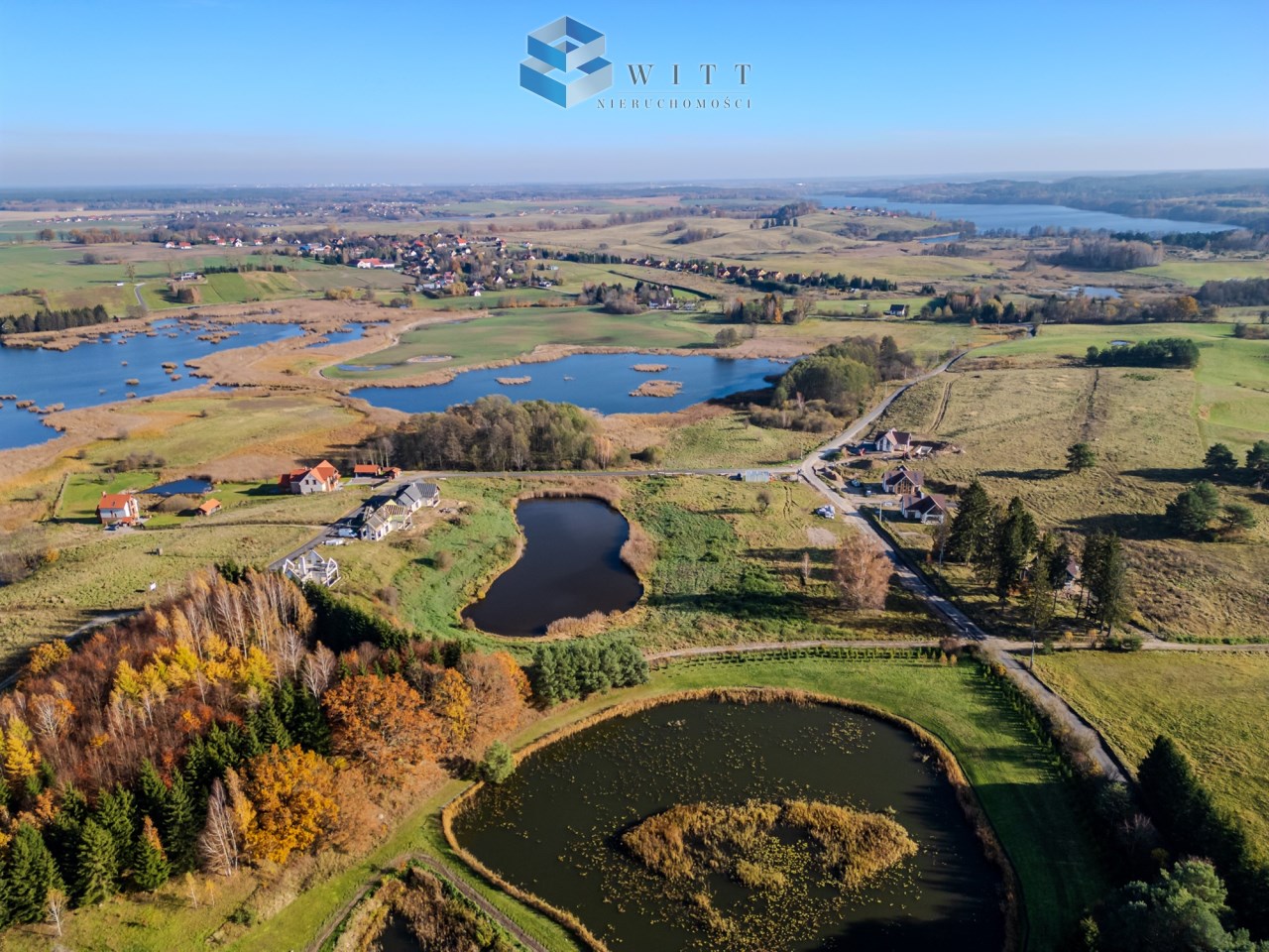 Widok z lotu ptaka na malowniczą wieś otoczoną wodami, z zielonymi polami i lasami w tle. Widać domy oraz tereny rekreacyjne, a także rozległe stawy.