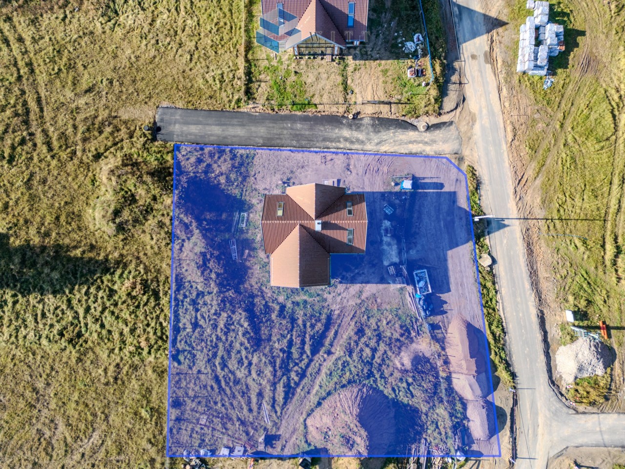 Widok z lotu ptaka na działkę budowlaną z nowym domem, otoczoną terenem z nieużytkami i drogą. Widać oznaczenie granic działki oraz materiały budowlane w pobliżu.