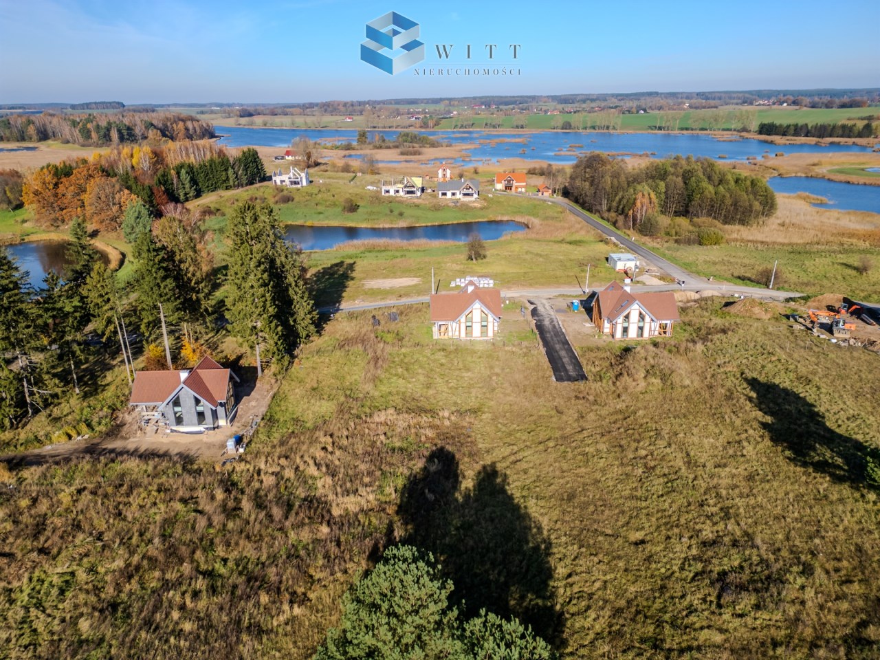 Widok z lotu ptaka na tereny rekreacyjne z domami w otoczeniu jezior i zieleni. W tle widać lasy i pola, a także nowo budowane nieruchomości.