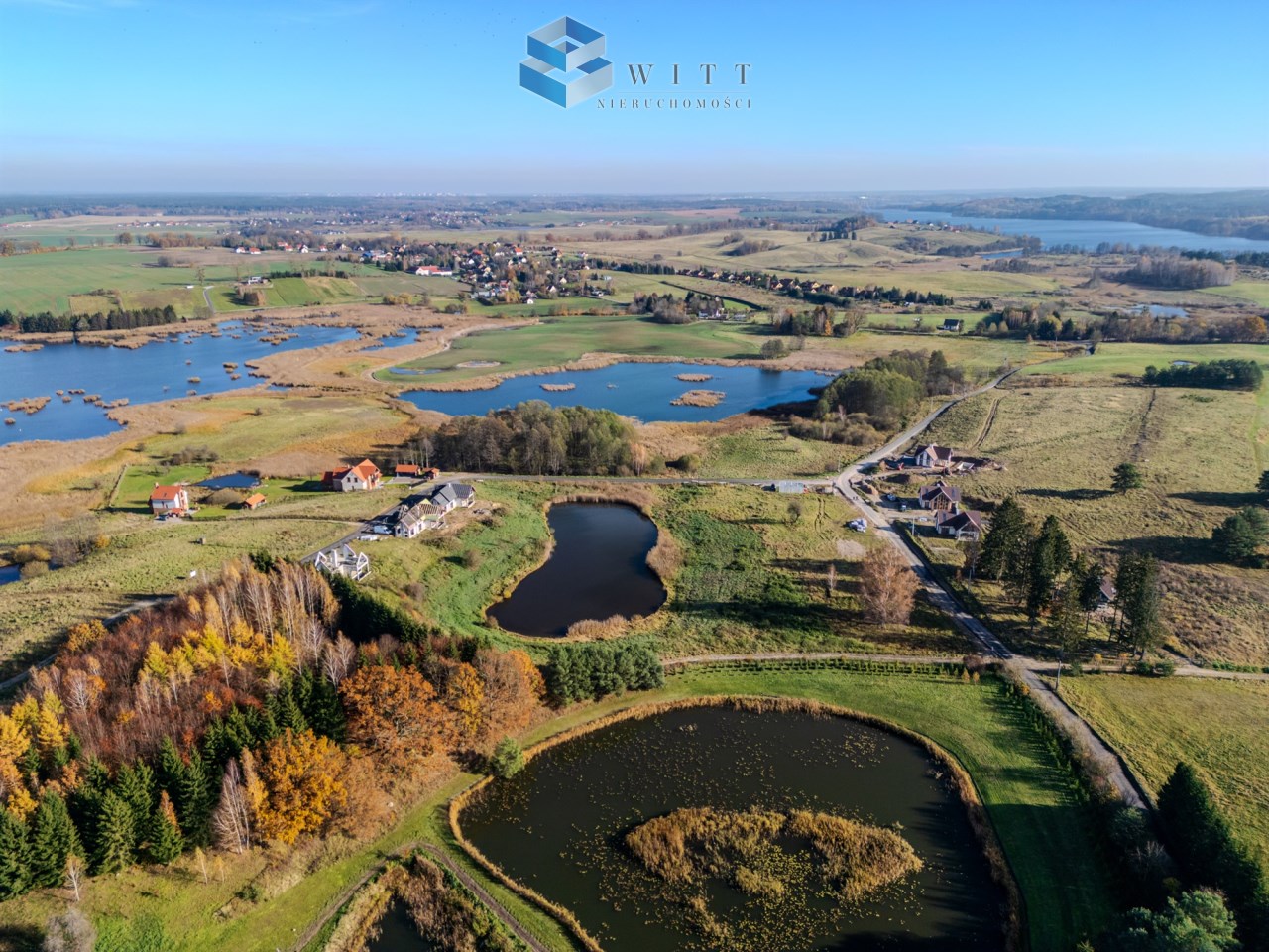 Widok z lotu ptaka na malowniczy krajobraz z jeziorami, zielonymi łąkami i domami w okolicy. W tle widać wieś oraz tereny podmokłe, otoczone drzewami w jesiennych kolorach.