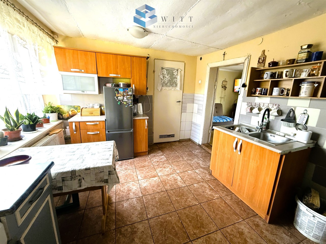 Kuchnia z drewnianymi meblami, nowoczesnym sprzętem AGD, w tym lodówką i zlewozmywakiem, oraz roślinami doniczkowymi na parapecie. W tle widoczna jasna ściana i drzwi prowadzące do innych pomieszczeń.