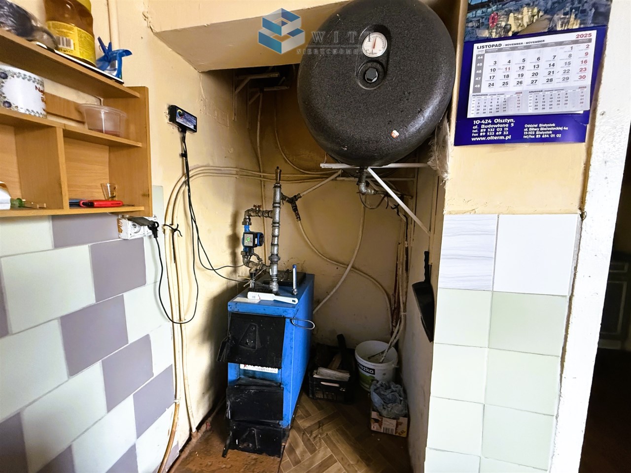 Instalacja grzewcza w kuchni, z niebieskim piecem, zbiornikiem na wodę oraz kalendarzem na ścianie.
