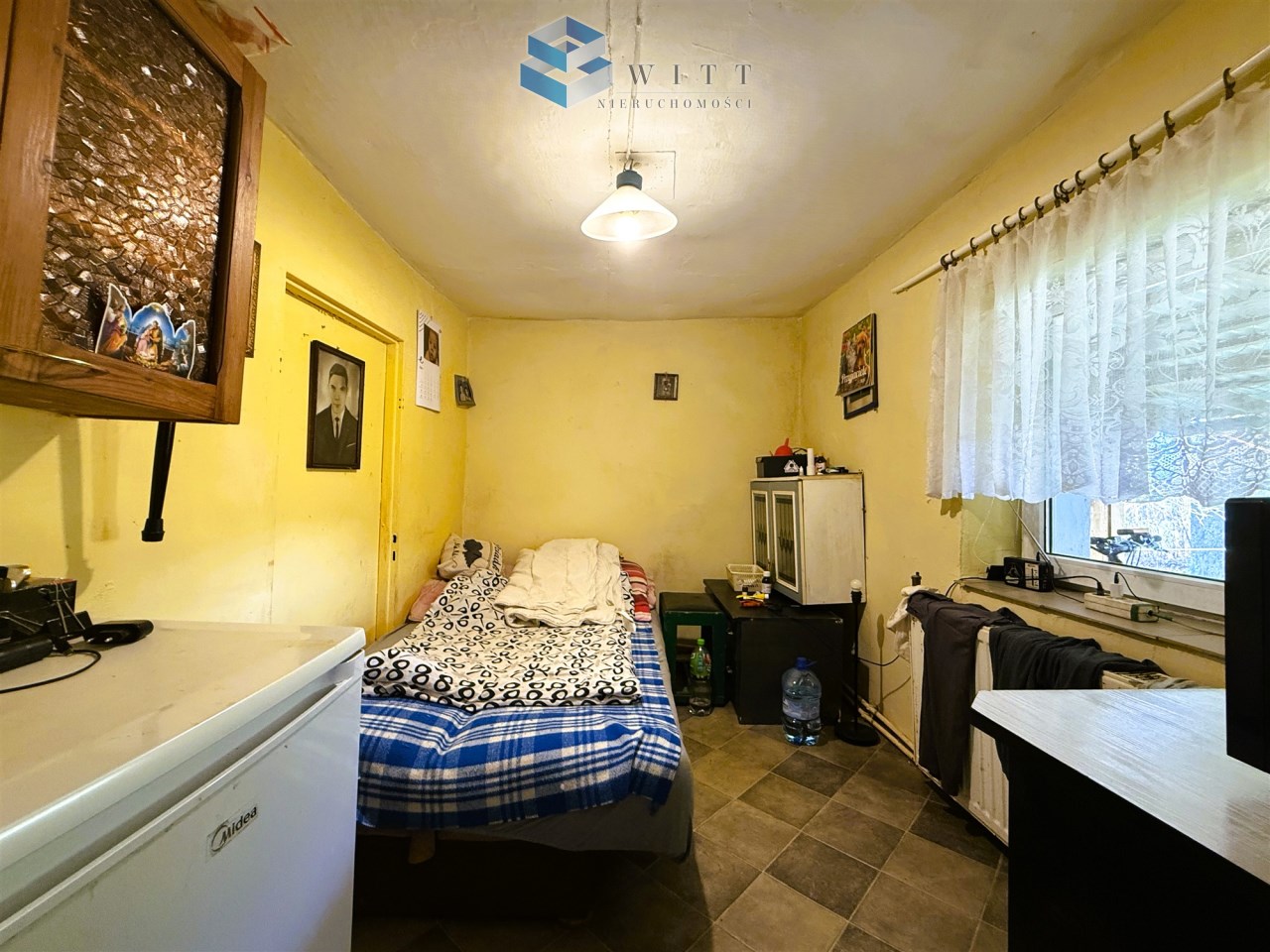 Wnętrze małego pokoju z żółtymi ścianami, łóżkiem z kocem w kolorowe wzory, biurkiem z elektroniką, lodówką i oknem z zasłoną. Na ścianach znajdują się zdjęcia i obrazy.