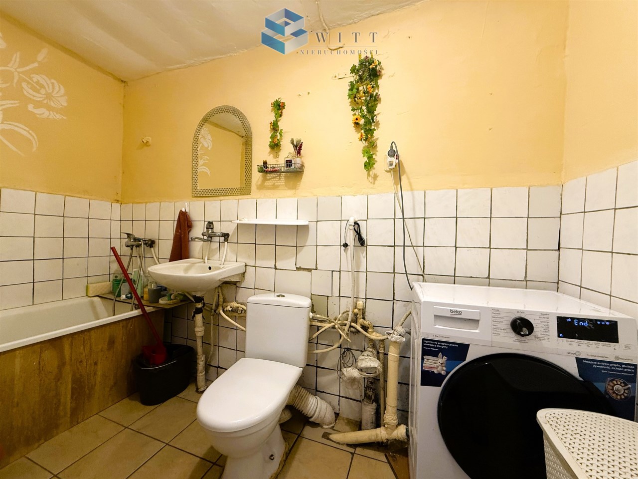 Łazienka z wanną, umywalką, toaletą i pralką, w jasnych kolorach, z białymi kafelkami na ścianach i podłodze, dekoracjami roślinnymi oraz akcesoriami do sprzątania.