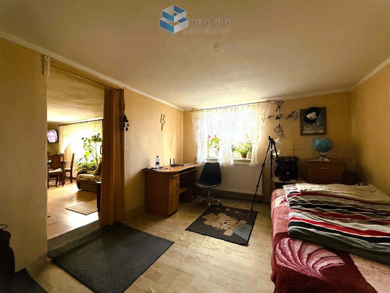 Wnętrze pokoju z łóżkiem, biurkiem i krzesłem, widok na sąsiedni pokój z roślinami i telewizorem.