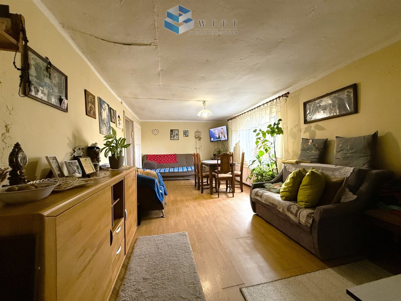 Zabytkowy salon z drewnianymi meblami, kanapą z poduszkami, stołem i krzesłami, oraz roślinami w doniczkach. Na ścianach obrazy i fotografie, a w tle telewizor. Oświetlony naturalnym światłem z okien.