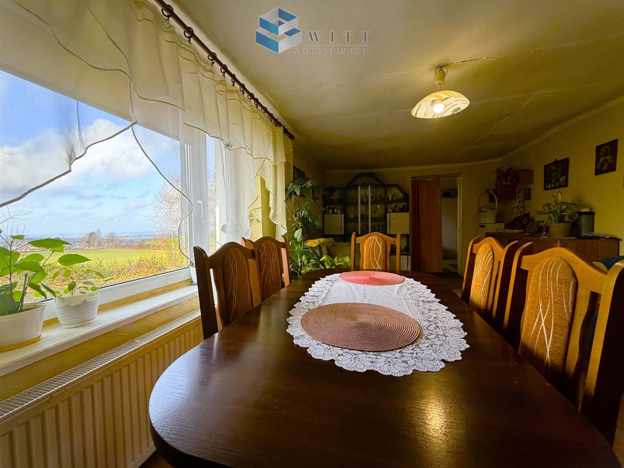 Widok na stół jadalny w jasnym wnętrzu, z roślinami na parapecie i dużym oknem, przez które widać krajobraz za domem.