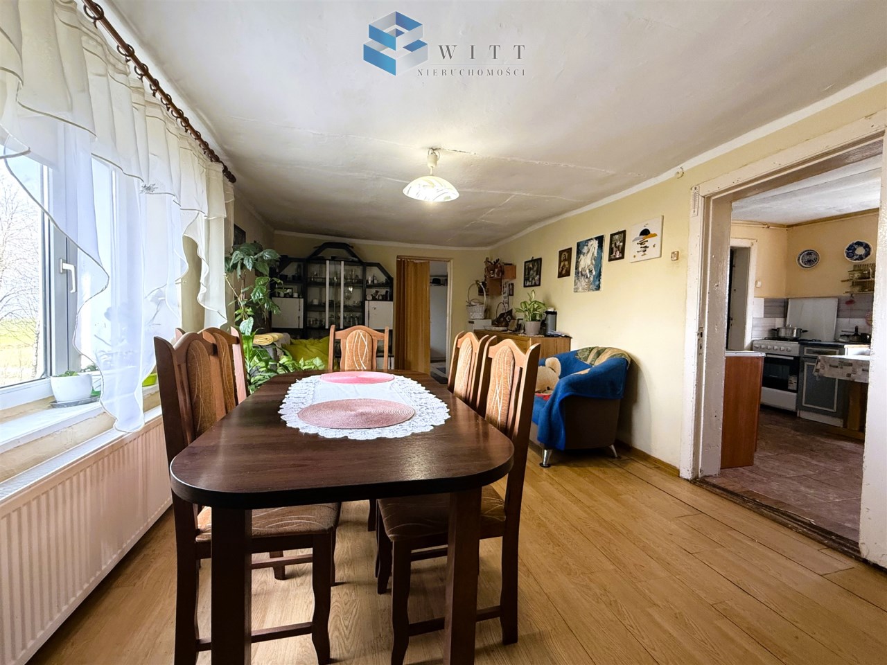 Widok na przytulną jadalnię z drewnianym stołem, krzesłami i zasłonami, w tle kuchnia oraz dekoracje na ścianach.