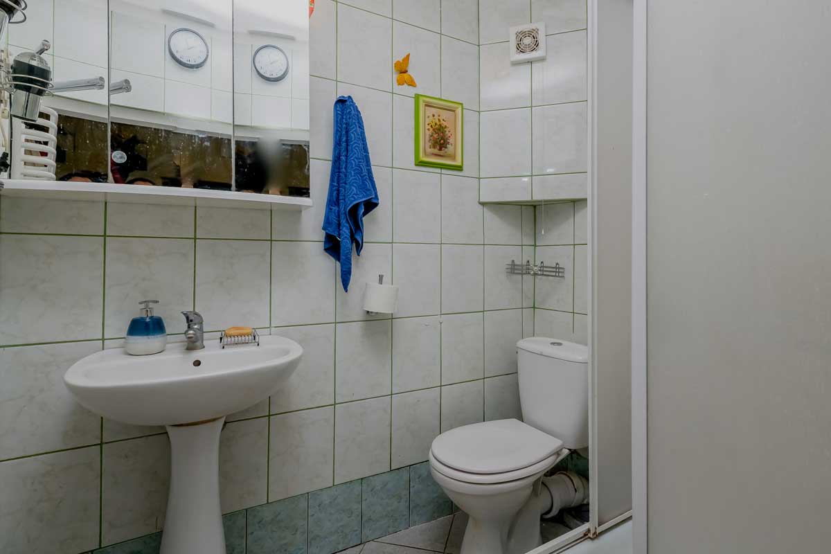 Łazienka z białymi kafelkami, umywalką, toaletą oraz lustrem. Na ścianie wisi niebieski ręcznik, a obok znajduje się dekoracyjny obrazek. W tle widoczne zegary.