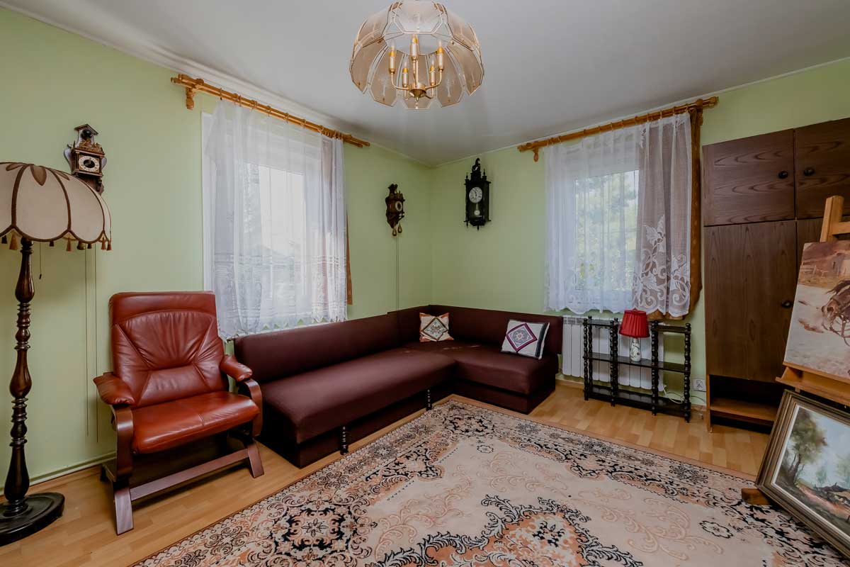 Wnętrze pokoju dziennego z zielonymi ścianami, brązową sofą w kształcie litery L, czerwonym fotelem, lampą stojącą oraz zegarem na ścianie. Na podłodze znajduje się dywan w stylu orientalnym, a okna są ozdobione białymi firankami.