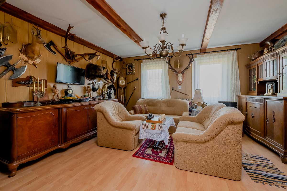 Stylowy salon z beżowymi fotelami, drewnianą komodą, telewizorem i dekoracjami w postaci trofeów myśliwskich na ścianach. W centralnej części stół z białym obrusem i szachami, a w tle jasne okna z firankami.