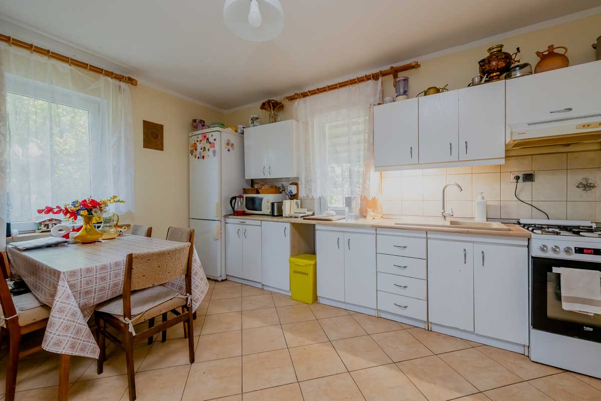 Jasna kuchnia z białymi meblami, stołem z krzesłami, oknem z firankami oraz sprzętem AGD, w tym lodówką i piekarnikiem. Na stole wazon z kolorowymi kwiatami.