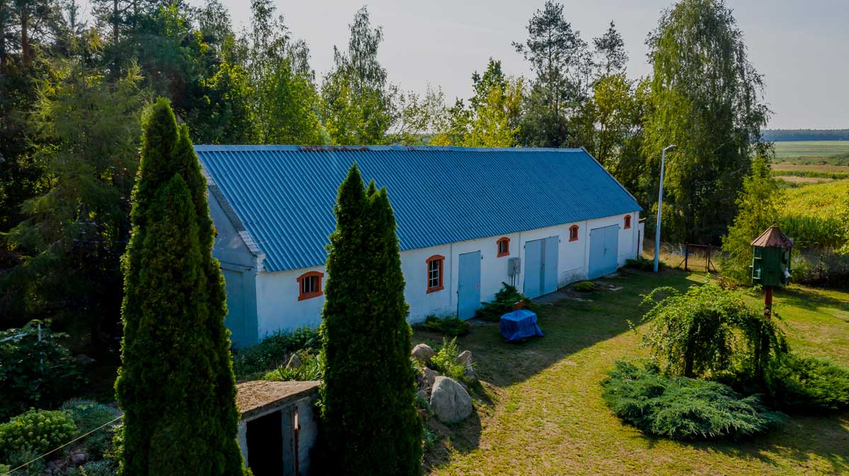 Widok na wiejski budynek z niebieskim dachem otoczony zielenią, drzewami i krzewami, na tle pola i jasnego nieba.