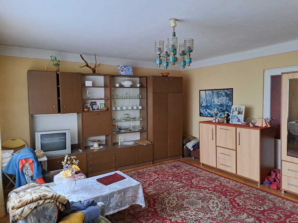 571014795_2735070643505872_7689226263220148475_n Wnętrze pokoju dziennego z meblami w stylu retro, kanapą, stołem z białym obrusem, telewizorem i dekoracjami na półkach. Na podłodze dywan w orientalny wzór.