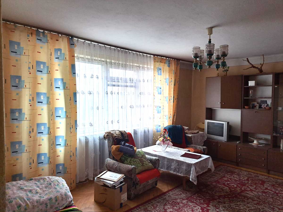 571091784_2735070650172538_5110875075263559824_n Zagospodarowane wnętrze salonu z zasłonami w kolorowe wzory, kanapami, stolikiem oraz telewizorem, oświetlone naturalnym światłem wpadającym przez okno.