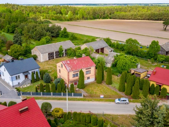 Widok z lotu ptaka na wiejską okolicę z domami jednorodzinnymi, ogrodami, polami uprawnymi i lasem w tle.
