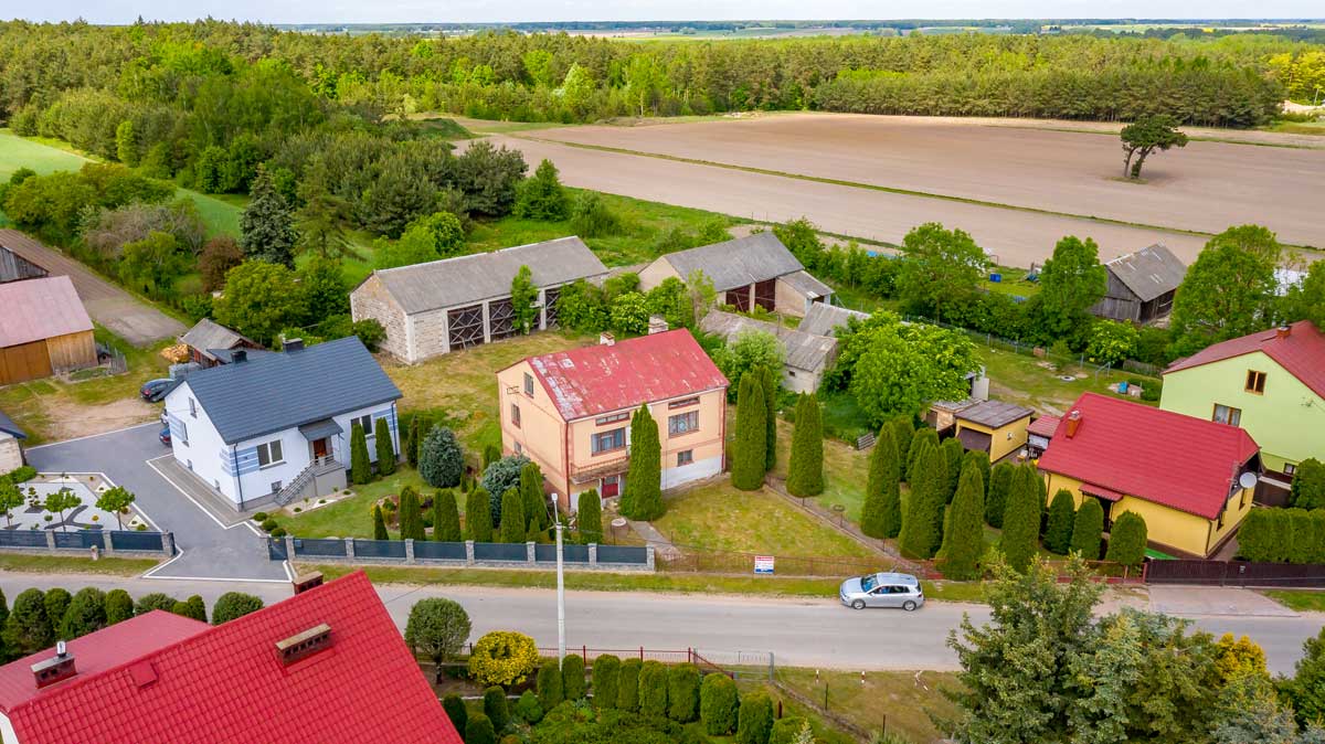 571109941_2735070886839181_5442151009193974120_n Widok z lotu ptaka na wiejską okolicę z domami jednorodzinnymi, ogrodami, polami uprawnymi i lasem w tle.