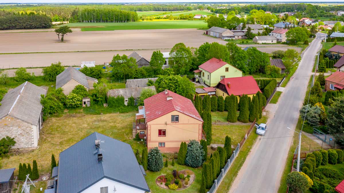 571130418_2735070923505844_5416789726225069786_n Widok z lotu ptaka na wiejską okolicę z domami, ogrodami i polami uprawnymi. Widać różnorodne budynki mieszkalne i gospodarcze otoczone zielenią oraz asfaltową drogę.