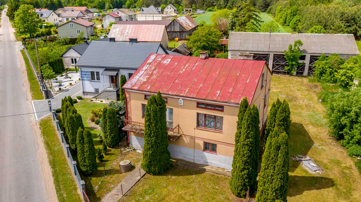 571136138_2735070830172520_1044421388222799258_n Widok na dom z czerwoną blachą na dachu, otoczony drzewami i innymi budynkami w wiejskiej okolicy. W tle widać zielone pola i drogi.