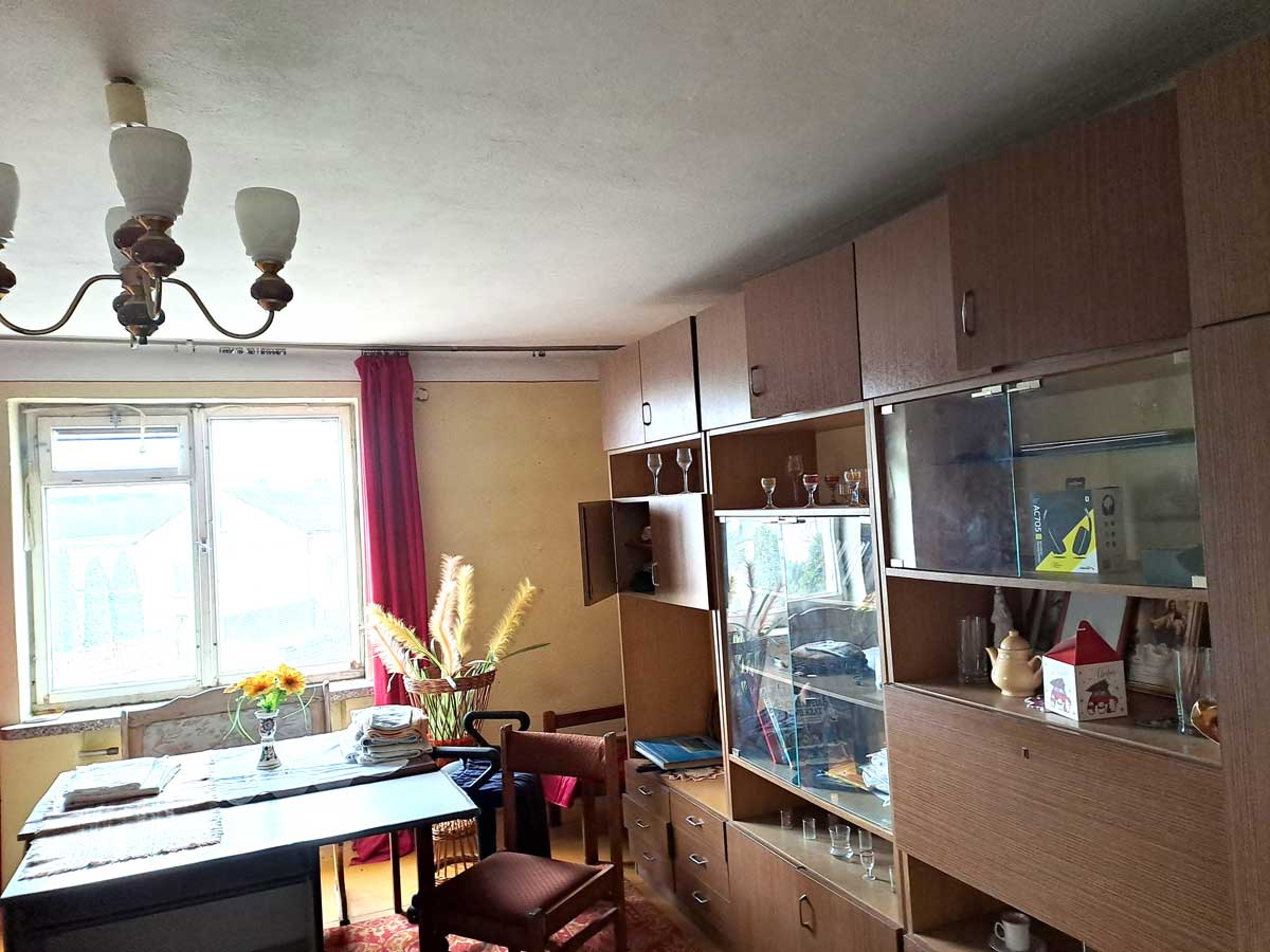 Wnętrze pokoju dziennego z drewnianymi meblami, stołem, krzesłami, dużym oknem z zasłonami oraz witryną z dekoracjami.