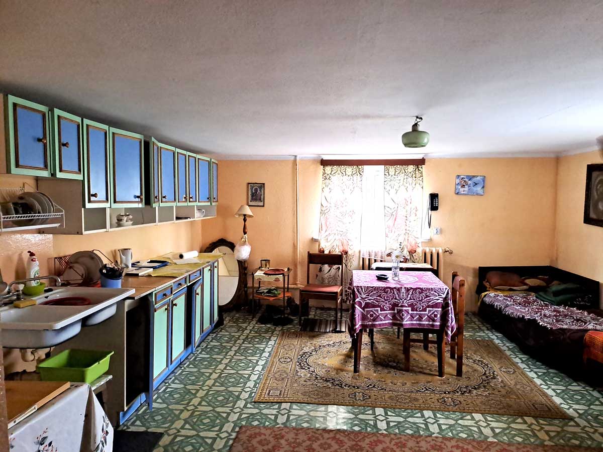 571209268_2735070790172524_5381862338686251042_n Widok na kuchnię z jadalnią w stylu retro, z niebieskimi szafkami, stołem z obrusami oraz kanapą. W tle okna z zasłonami i lampą stojącą. Podłoga w wzory.