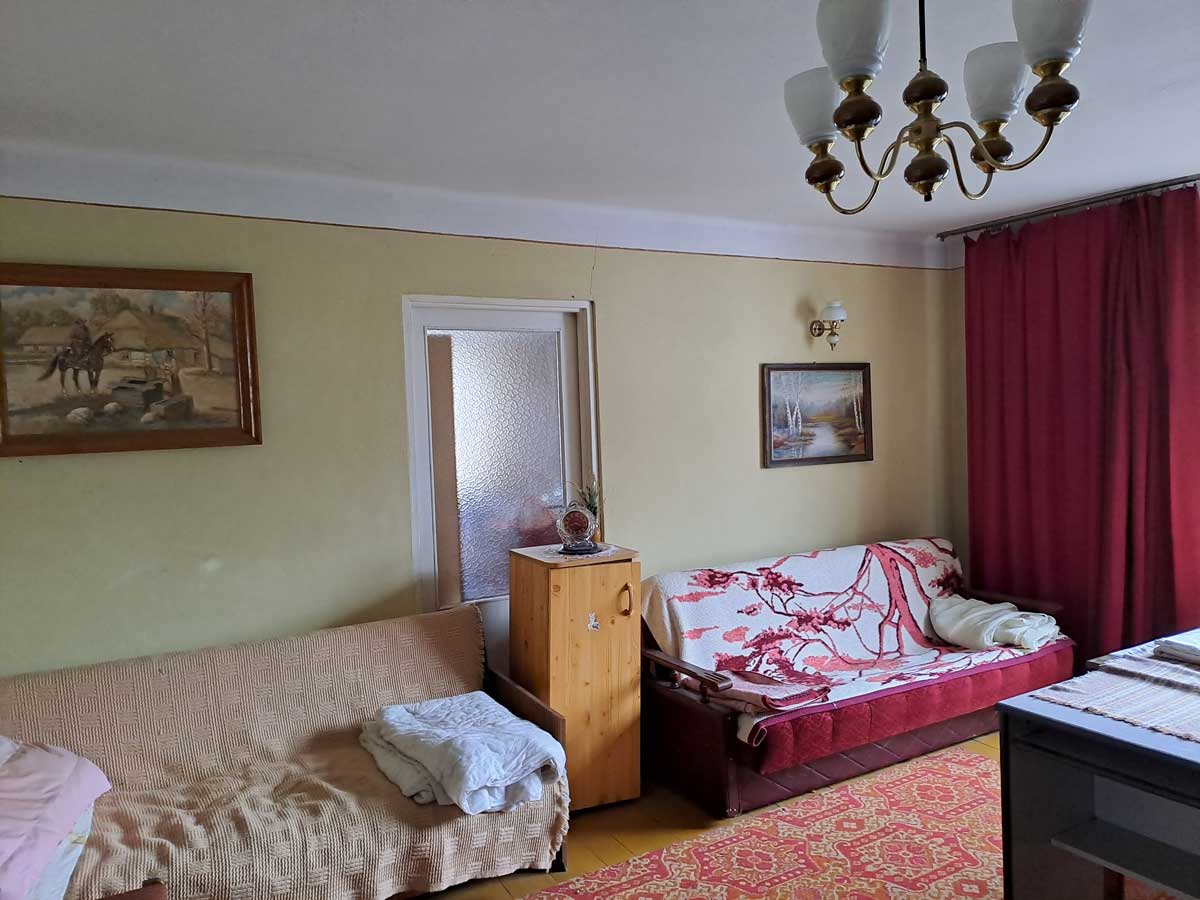 571216606_2735070716839198_1531556044560375195_n Zamknięta przestrzeń dzienna z dwoma kanapami, drewnianym stolikiem, lampą sufitową i obrazami na ścianach. Na podłodze dywan w kolorze czerwonym. Czerwone zasłony przy oknie.