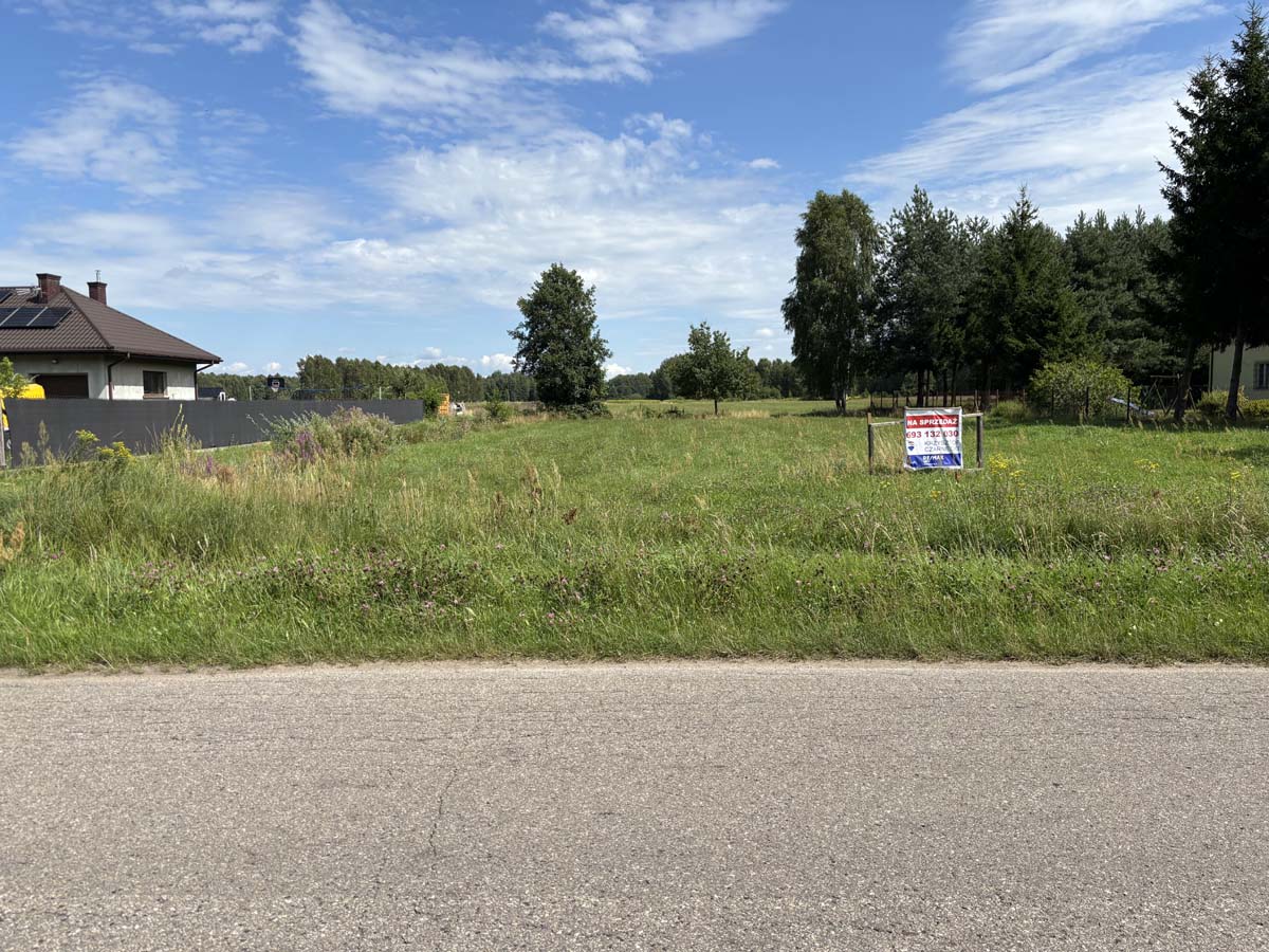 Widok na działkę na sprzedaż z oznaczeniem, w otoczeniu zieleni, z domem w tle i niebieskim niebem.