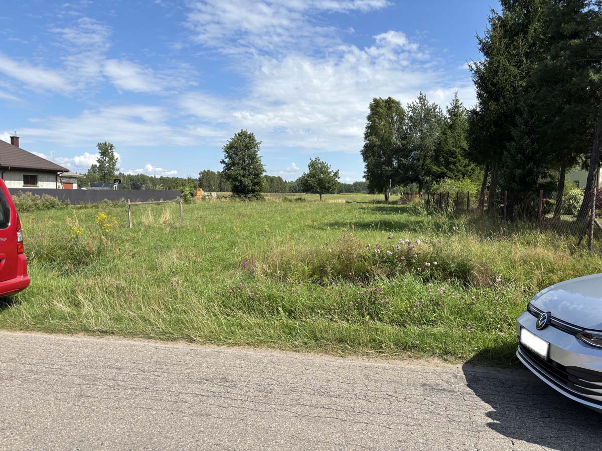 Widok na wiejską okolicę z zielonym polem, drzewami i dwoma samochodami zaparkowanymi przy drodze. W tle widać dom oraz ogrodzenie. Niebieskie niebo z białymi chmurami.