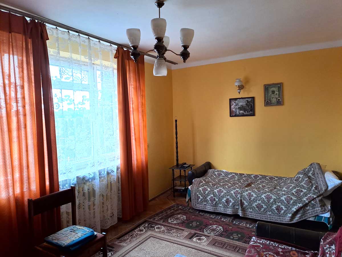 571363084_2735070646839205_6807544800182243058_n Wnętrze pokoju z żółtymi ścianami, dużym oknem z białymi firanami i pomarańczowymi zasłonami, kanapą z narzutą, dywanem oraz drewnianym krzesłem. Na ścianach znajdują się obrazy, a sufit zdobi lampa z kilkoma kloszami.