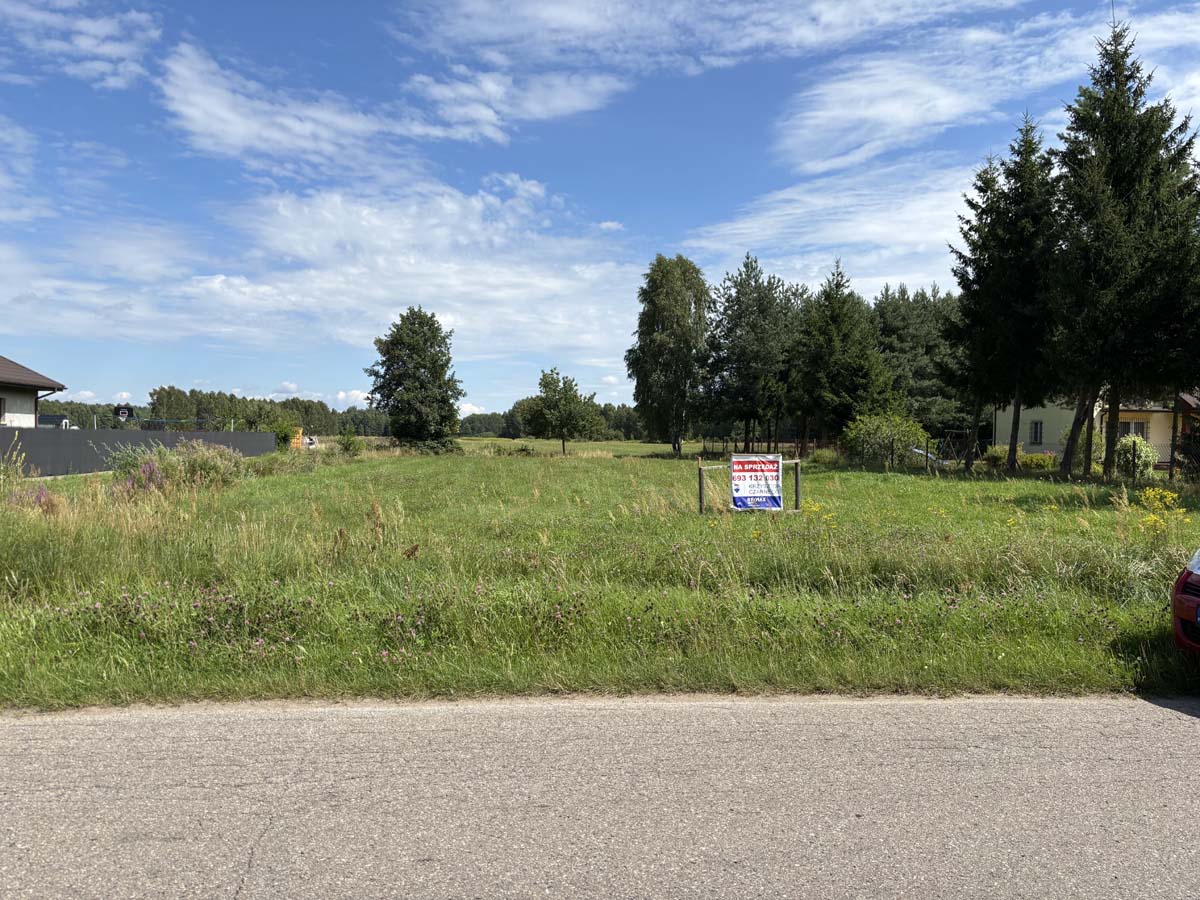 Widok na działkę budowlaną z ogłoszeniem "Na sprzedaż" w otoczeniu zieleni, drzew i nieba z chmurami.