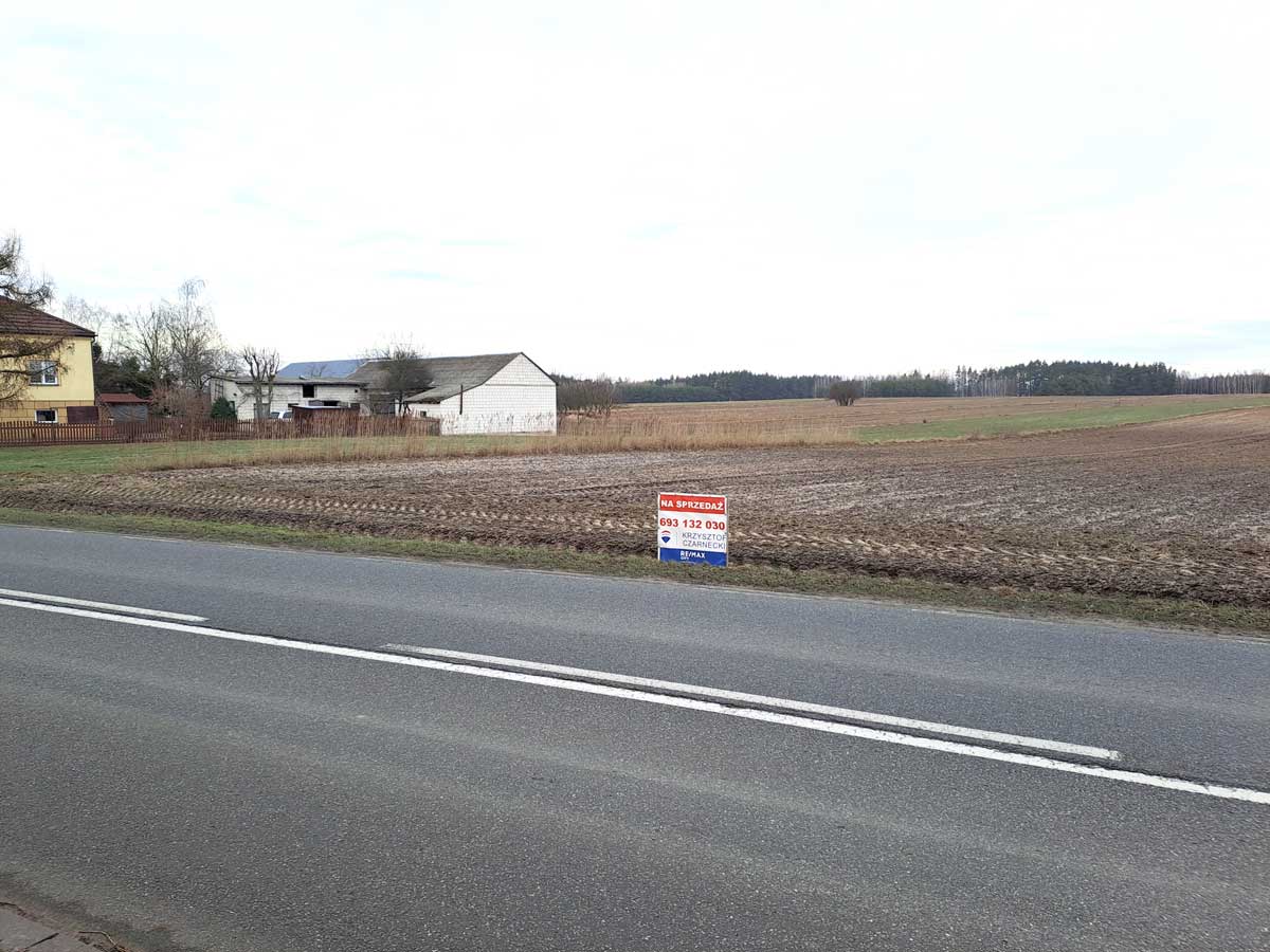 Widok na wiejską drogę z tablicą ogłoszeniową "Na sprzedaż" oraz widocznymi zabudowaniami i polami uprawnymi w tle.