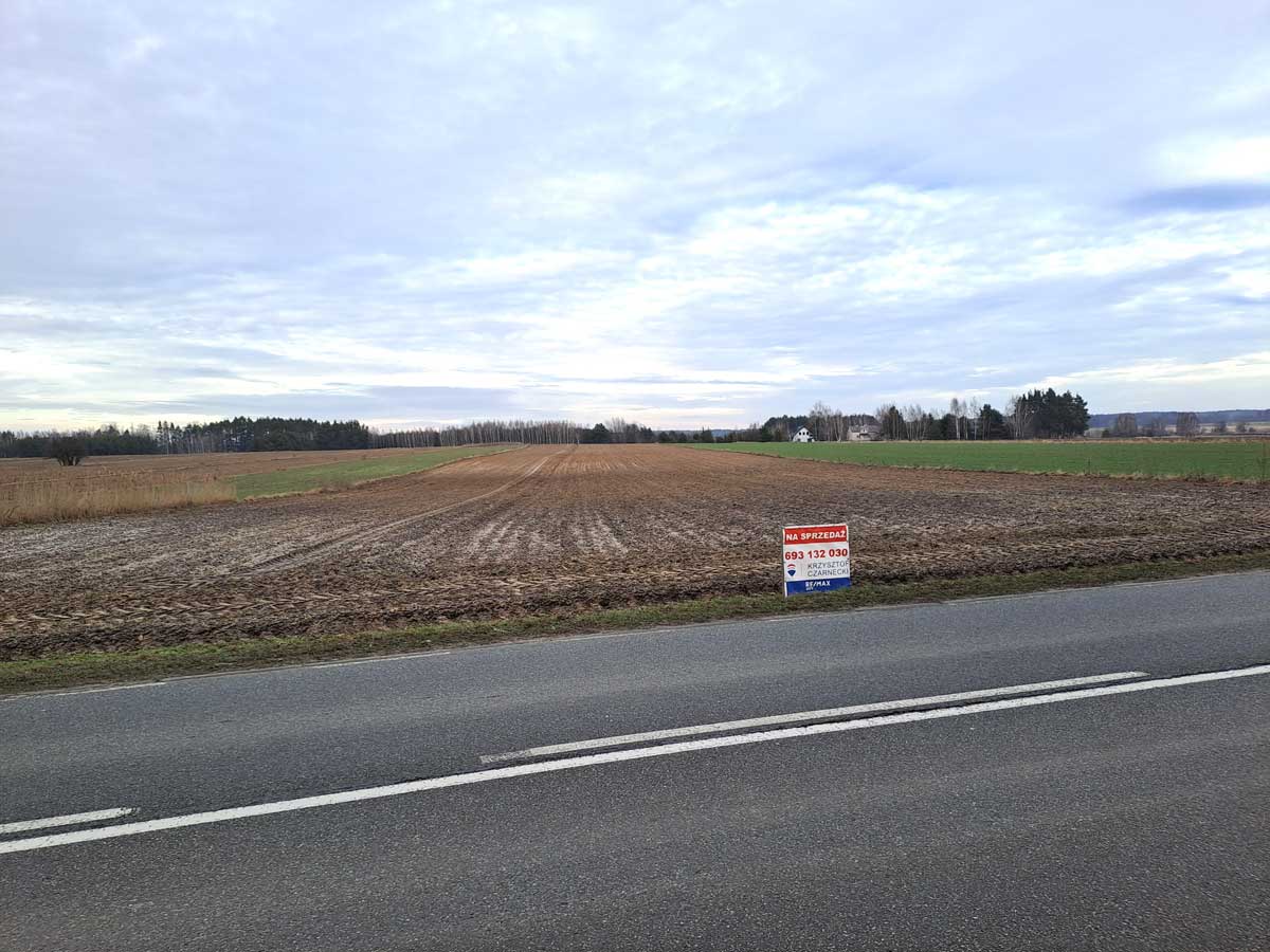 577034891_2747697488909854_7448285859560371558_n Widok na działkę na sprzedaż przy drodze, z oznaczeniem kontaktowym. W tle pola uprawne i zadrzewienia.