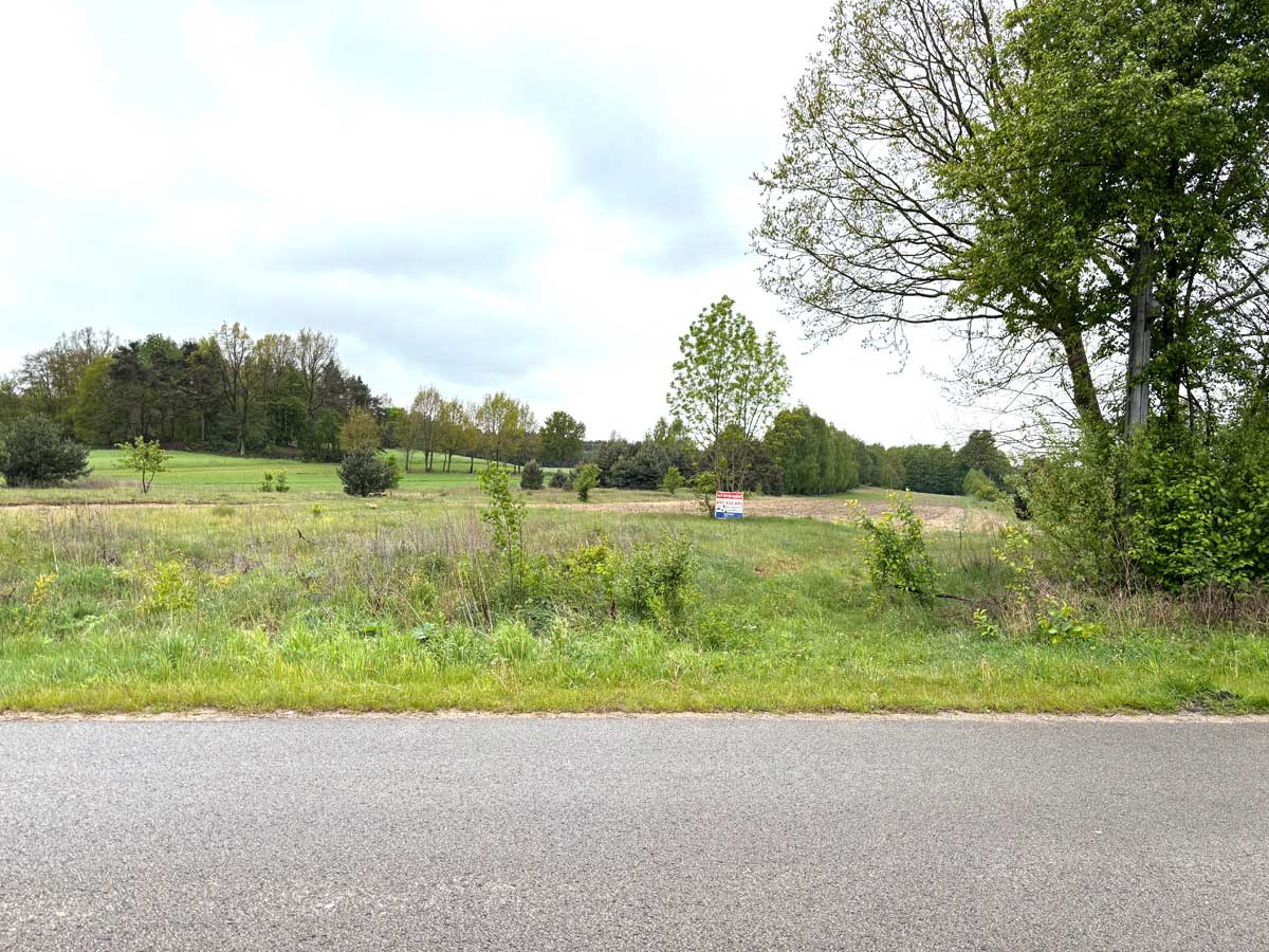 587253461_2764824130530523_2337932724051120281_n Widok na zieloną łąkę z drzewami i krzewami, w tle widoczne niewielkie wzgórza oraz znak informacyjny.